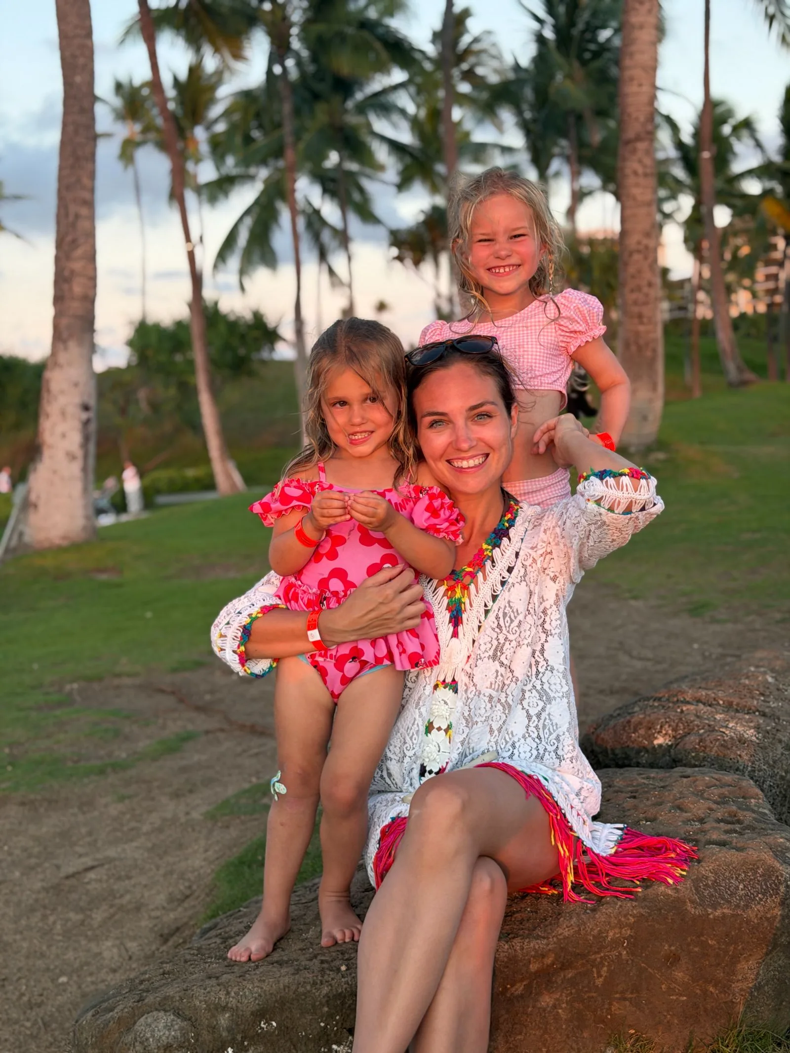 Kiersten sitting on a lava rock at dusk in a white lace cover-up, one daughter on her lap and the other perched on her shoulders, coconut palms and a pink sunset sky behind them
