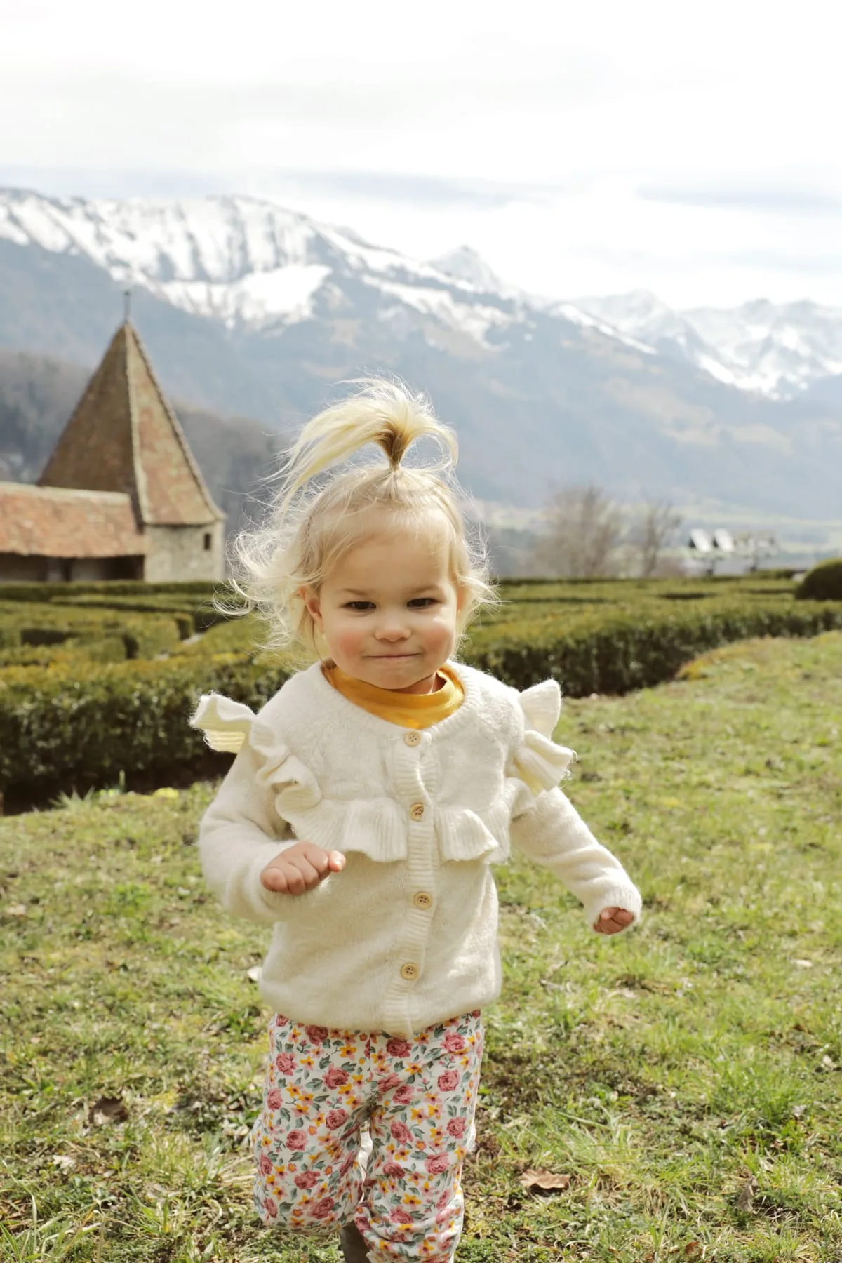 A toddler in a cream ruffled sweater standing in a field below snowy mountains
